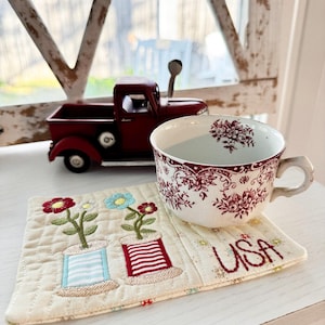 May include: A burgundy toy truck and a floral teacup rest on a white surface. A quilted coaster with embroidered flowers and the word "USA" is under the teacup. The teacup has a floral pattern.