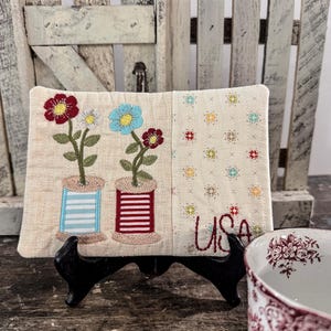May include: A rectangular, quilted fabric piece with embroidered flowers in red, blue, and white, atop thread spools. The word "USA" is embroidered in red. The piece is displayed on a black stand, with a floral-patterned bowl in the foreground.
