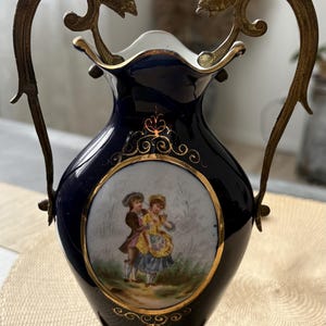 Może przedstawiać: Ciemnoniebieski wazon porcelanowy z centralnym owalnym medalionem przedstawiającym malowaną scenę z dwiema postaciami w strojach z epoki. Wazon posiada ozdobne złote metalowe uchwyty oraz pasującą podstawę z misternymi zdobieniami.