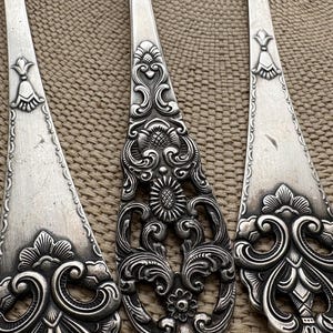 Può includere: Primo piano di posate antiche, tra cui un cucchiaio e forchette, con elaborati disegni floreali e a volute. Gli utensili d'argento sono disposti su una superficie strutturata dai toni neutri, che mostra gli intricati dettagli delle posate vintage.