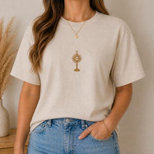 May include: Beige short-sleeved t-shirt with a gold-colored religious symbol on the front. The symbol is a detailed monstrance, a vessel used in the Catholic Church to display the Eucharist. The shirt is paired with blue jeans.