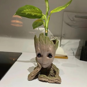 May include: A Groot-shaped planter holds a green plant with variegated leaves. The planter is a detailed sculpture with a textured, wood-like appearance, featuring large black eyes and a gentle smile. The plant's stem rises from the planter's head.
