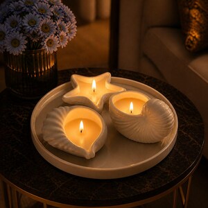 May include: Three lit candles in seashell and starfish shapes on a white tray. The candles are on a dark marble-topped table with a gold base. A vase of blue flowers is in the background.