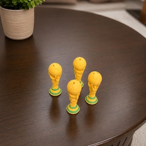 May include: Four small yellow and green figurines shaped like the FIFA World Cup trophy. Each figurine features a spherical top section supported by a base with a green ring detail. The items are arranged in a cluster on a dark brown surface.