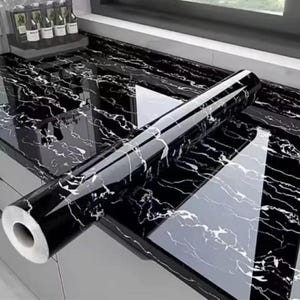 May include: A roll of black marble-effect contact paper rests on a matching countertop. The glossy surface reflects light, displaying white veining against the black. The countertop and paper create a cohesive, modern appearance. The roll is approximately 30 cm in diameter.