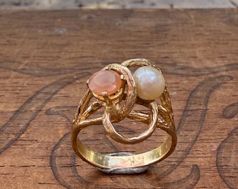 Può includere: Un anello d'oro con una pietra preziosa color pesca e una perla bianca. L'anello ha un design intricato e intrecciato. L'anello è posto su una superficie di legno con il testo "NASSAU ST." e "OUGHBY ST."