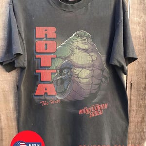 May include: Dark gray t-shirt featuring a graphic of a large, green alien character with the text "ROTTA" in red block letters. Below the graphic are the words "The Hutt" and "MANDALORIAN GROGU". The shirt is labeled "COMFORT COLORS" and "PEPPER".