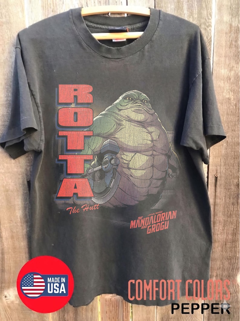 May include: Dark gray t-shirt featuring a graphic of a large, green alien character with the text "ROTTA" in red block letters. Below the graphic are the words "The Hutt" and "MANDALORIAN GROGU". The shirt is labeled "COMFORT COLORS" and "PEPPER".