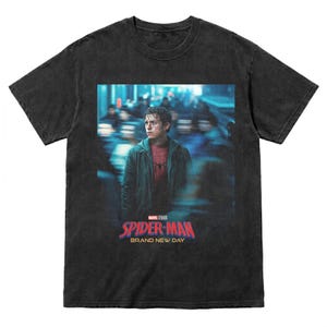 Pode incluir: T-shirt cinzento escuro com um gr&aacute;fico de cartaz de filme. O gr&aacute;fico inclui as palavras "Spider-Man" em vermelho e o slogan "Brand New Day". A imagem na camisa mostra uma pessoa com uma jaqueta verde.