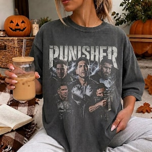 May include: A charcoal grey short-sleeve t-shirt featuring a graphic print of The Punisher with multiple character portraits and lightning effects. The text THE PUNISHER is printed in a distressed white font across the upper chest area.