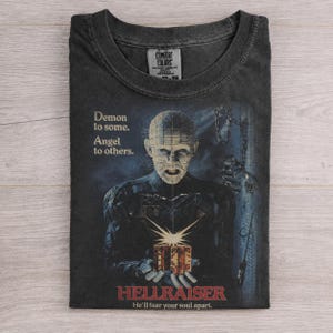 May include: A charcoal gray crew neck t-shirt featuring a graphic of Pinhead from Hellraiser holding a puzzle box. The text reads: Demon to some. Angel to others. HELLRAISER He'll tear your soul apart. The shirt has a Comfort Colors brand label.