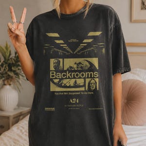 May include: A charcoal grey short-sleeve t-shirt featuring a graphic print for the movie Backrooms. The design includes yellow text, multiple film stills, the A24 logo, and the phrase You Are Not Supposed To Be Here, with a vintage washed finish.
