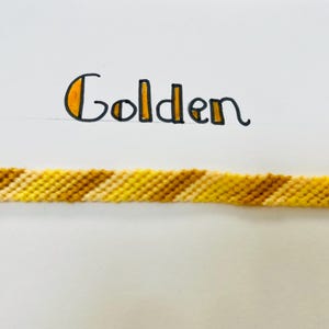 May include: A woven friendship bracelet in shades of yellow and brown. The word "Golden" is written above the bracelet in black ink with yellow accents. The bracelet is displayed on a white background.