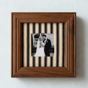 May include: A square wooden picture frame with a dark brown finish. The inner matting features a vertical black and beige striped fabric pattern. Centered within the frame is a black and white photograph of a couple in wedding attire.