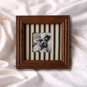 May include: Square wooden picture frame with a dark brown finish. The frame has a striped fabric insert in black and cream, surrounding a black and white photograph of a person and a dog. The frame is on a white surface.