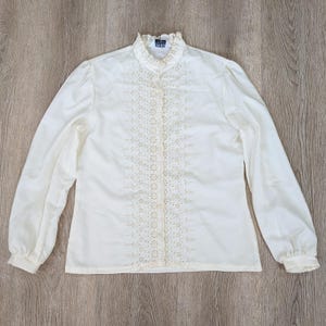 May include: A cream-colored long-sleeve blouse featuring a ruffled high collar and a central vertical panel of intricate eyelet embroidery. The garment includes long sleeves with gathered cuffs and a button-down front closure with a visible brand label.