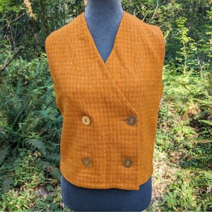 May include: A double-breasted vest in a textured mustard yellow fabric with a subtle white grid pattern. The garment features a V-neckline, structured shoulders, and four round, light-colored buttons arranged in two vertical rows on the front.