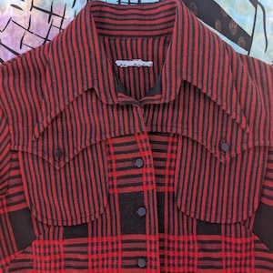 May include: Red and black plaid shirt with black accents. The shirt features a collared neckline, two front pockets, and black buttons. The fabric has a vertical striped pattern on the upper portion and a plaid pattern on the lower portion.