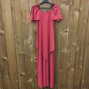 May include: A dusty rose-colored maxi dress with short, draped sleeves and a unique layered design. The dress is hanging on a black hanger against a wooden plank background. The dress has a V-neckline and a flowing skirt.
