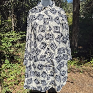 May include: A long-sleeve button-down shirt featuring a repeating pattern of dark gray square motifs on a light cream background. The shirt has a standard collar, a full-length front button placket with light-colored buttons, and a straight hemline.