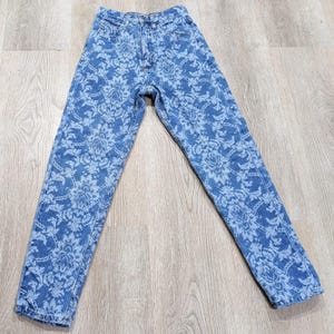 May include: High-waisted denim jeans in a medium blue wash featuring an all-over white floral damask pattern. These pants include a standard button and zipper fly closure, five pockets, and a straight-leg cut with visible stitching throughout.