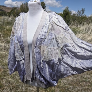 May include: A patchwork cardigan in shades of lavender, gray, and cream, displayed on a mannequin. The open-front design features a ribbed collar and cuffs, with various fabric textures and patterns, including paisley and lace details. The sleeves are wide and flowing.