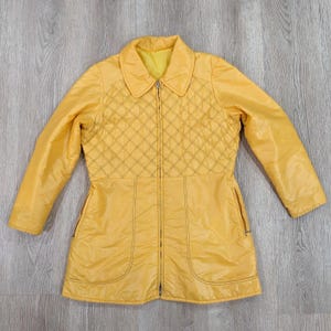 May include: A vintage yellow jacket with a quilted diamond pattern on the upper chest. The jacket has a collared neckline, long sleeves, and a full-length zipper. Two pockets are visible on the lower front. The jacket is made of a shiny, possibly nylon, material.