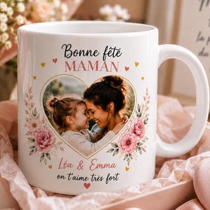 Peut inclure: Un mug en céramique blanc orné d'une photo en forme de cœur d'une femme et d'un enfant, entourée d'illustrations florales roses. Le texte indique Bonne fête MAMAN, Léa & Emma on t'aime très fort, avec des cœurs dorés et roses.