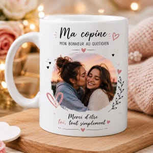 Puede incluir: Una taza de cerámica blanca con una foto circular de dos personas abrazándose. El texto dice "Ma copine", "MON BONHEUR AU QUOTIDIEN" y "Merci d'être toi, tout simplement". El diseño incluye pequeños corazones rosas, corazones negros y hojas.