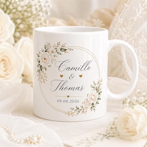 Può includere: Una tazza in ceramica bianca con una cornice circolare dorata decorata con rose bianche e fogliame verde. Il testo riporta Camille & Thomas con la data 08.08.2026 centrata sotto. Piccoli cuori dorati sono posizionati tra i nomi e la data.