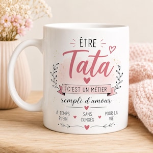 Puede incluir: Una taza de cerámica blanca con el texto en francés "ÊTRE TATA C'EST UN MÉTIER REMPLI D'AMOUR À TEMPS PLEIN SANS CONGÉS POUR LA VIE" en letras negras y rosas, decorada con pequeños corazones rosas e ilustraciones de hojas negras.