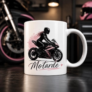 Puede incluir: Una taza de cerámica blanca con una ilustración gráfica de una persona con equipo negro sobre una motocicleta deportiva negra y rosa. Detrás hay una mancha de acuarela circular de color rosa suave. La palabra Motarde aparece en letra cursiva.