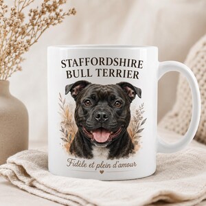 Può includere: Una tazza in ceramica bianca con un'illustrazione dettagliata di un Staffordshire Bull Terrier nero. Il testo "STAFFORDSHIRE BULL TERRIER" è stampato sopra il cane, e "Fidèle et plein d'amour" è scritto sotto con una piccola icona a cuore.