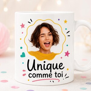 Può includere: Una tazza in ceramica bianca con una cornice circolare centrale contenente la fotografia di una persona che fa l'occhiolino e sorride. La tazza è decorata con stelle, punti e cuori colorati e include il testo nero "Unique comme toi".