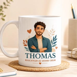 Può includere: Una tazza in ceramica bianca con un'illustrazione in stile cartoon di un uomo con capelli scuri e barba, che indossa una camicia verde acqua. Il testo "THOMAS" è scritto in blu scuro, seguito da "ÉDUCATEUR AU GRAND CŒUR" in arancione, con cuori e foglie.