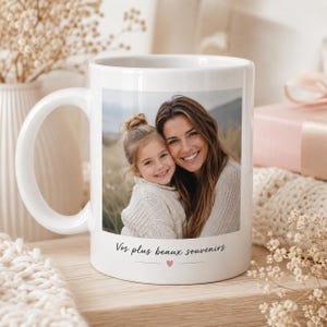 Puede incluir: Una taza de cerámica blanca con una impresión fotográfica cuadrada de un adulto y un niño sonrientes. Debajo de la imagen, el texto "Vos plus beaux souvenirs" aparece en una fuente cursiva negra, junto a un pequeño corazón rosa.