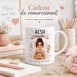 Pode incluir: Uma caneca de cerâmica branca com uma ilustração de desenho animado de uma pessoa de cabelo castanho e óculos segurando livros. A caneca exibe o texto AESH, Camille e Merci pour ton aide précieuse, rodeado por corações rosa e folhas.