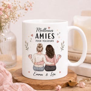 Pode incluir: Uma caneca de cerâmica branca com uma ilustração de duas pessoas abraçadas segurando canecas. O texto "Meilleures AMIES POUR TOUJOURS" e "Emma & Léa" está escrito em letras pretas, decorado com pequenos corações e estrelas cor-de-rosa.