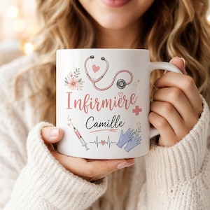 Pode incluir: Uma caneca de cerâmica branca com um estetoscópio rosa, uma seringa, luvas médicas azuis e detalhes florais. O texto diz "Infirmière" em letra rosa e "Camille" em cursivo preto, acompanhado por uma linha de batimento cardíaco e corações.