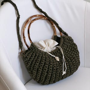 May include: A dark olive green crocheted handbag featuring a textured ribbed pattern, two round bamboo handles, and a long braided shoulder strap. The bag includes an off-white fabric drawstring closure with small white beads at the ends of the cords.