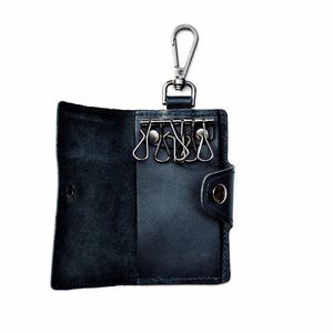 May include: Black leather key holder with a silver carabiner clip. The key holder is open, showing key rings. It has a snap closure and is designed to hold multiple keys. The background is white.