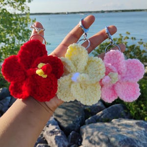 May include: Three crochet flower keychains in red, pale yellow, and light pink. Each flower features five rounded petals and a central stamen, attached to a silver-toned metal split ring and a lobster claw clasp for secure attachment to bags or keys.