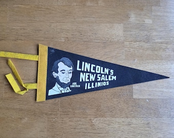 May include: Black felt pennant with yellow trim and a yellow ribbon. The pennant features a white illustration of Abraham Lincoln and the text "LINCOLN'S NEW SALEM ILLINOIS". A vintage souvenir.