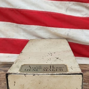 May include: A white metal drawer with a black handle. The drawer has a gold label that reads "The American Home Menu Maker".
