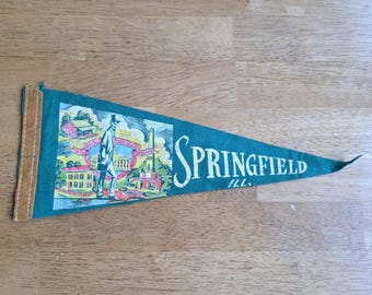 May include: A vintage green pennant with the word "Springfield" in white script. The pennant features an illustration of a man and buildings. The pennant is trimmed with brown fabric and has a pointed end.