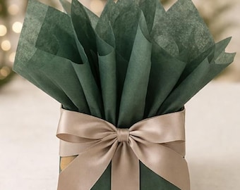 May include: A wrapped gift box with dark green paper and a beige satin ribbon tied in a bow. The top of the box is filled with dark green tissue paper, fanned out to create a decorative effect. The background is blurred with soft, out-of-focus lights.