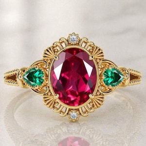 May include: A gold ring featuring a large oval-cut red center stone, flanked by two pear-cut green side stones. The setting is detailed with intricate metalwork, including small clear accent stones at the top and bottom of the oval center.