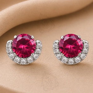 May include: A pair of stud earrings featuring a large, round, vibrant red center stone set in silver-toned metal. Each earring is accented with a semi-circular halo of small, clear, sparkling stones along the bottom edge of the setting.