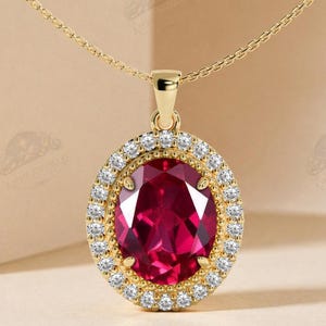 May include: A gold necklace featuring an oval-shaped pendant with a large, deep red gemstone at the center. The red stone is surrounded by a halo of small, clear, round-cut white stones set in a gold frame, suspended from a delicate gold chain.
