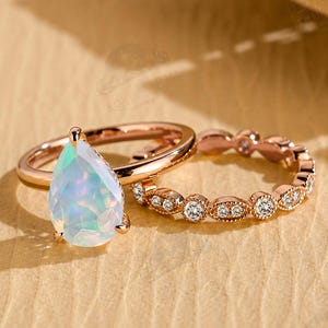 May include: A rose gold ring set featuring a pear-shaped opal center stone with iridescent flashes of color and a matching band adorned with a series of small, round, clear stones set in a delicate, textured metal frame with milgrain detailing.